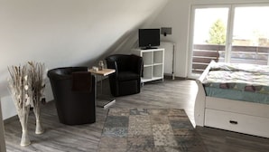 Ein modernes, kompaktes Zimmer mit schrägverdeckter Decke, einem Bett, einem kleinen Schreibtisch, einem Sessel und einem Fernseher.