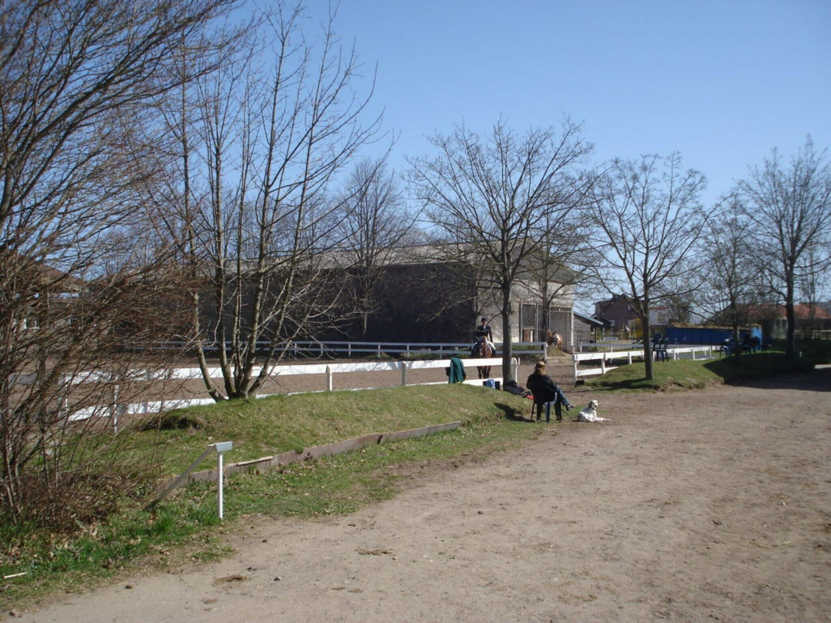 A fenced outdoor area with a dirt path, trees, and a building in the background.
