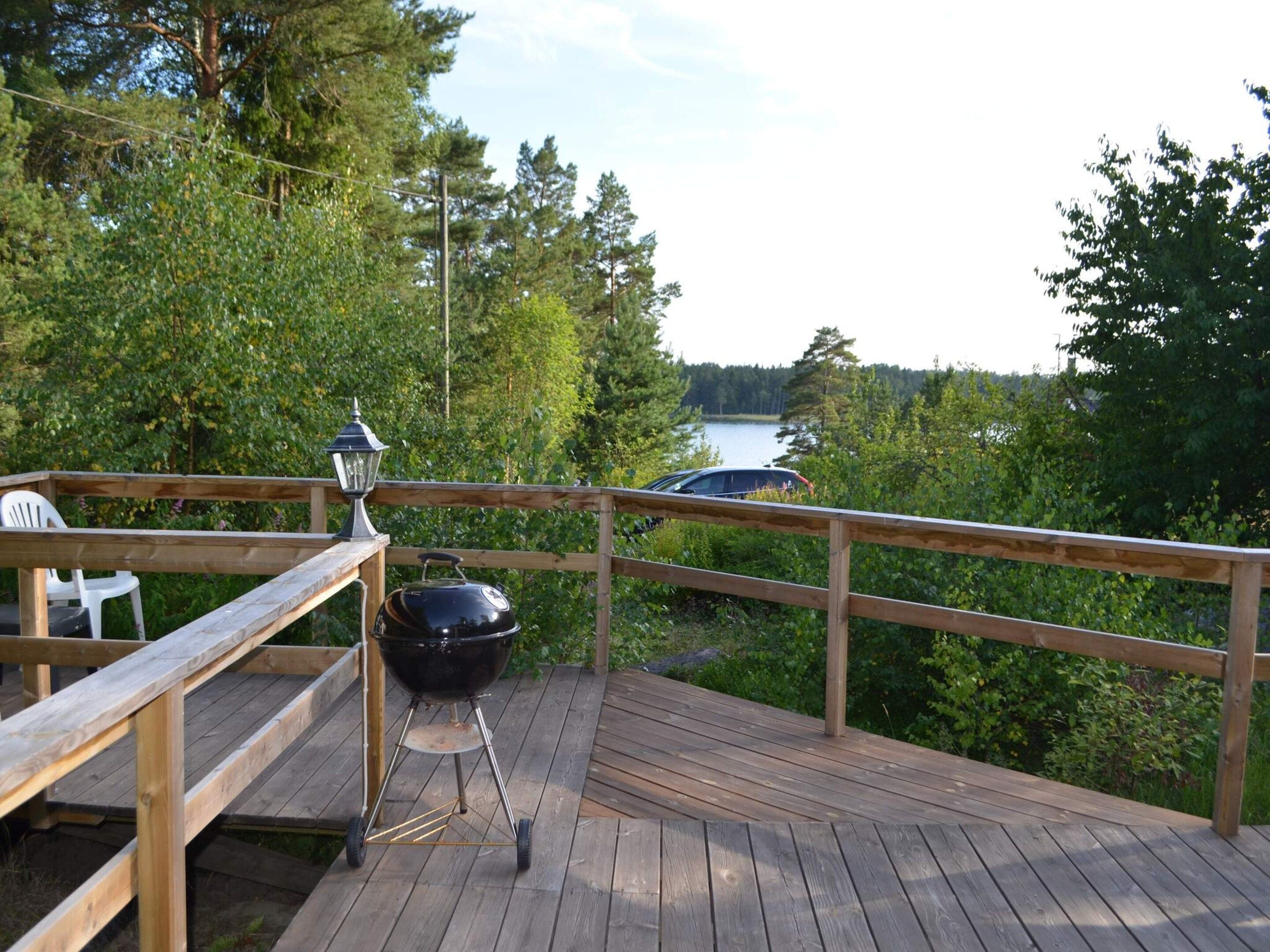 A wooden deck with a barbecue grill and a lantern, overlooking a lake surrounded by trees.