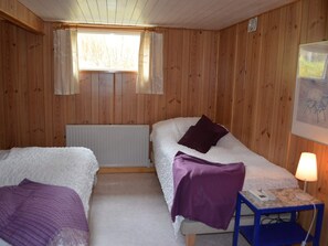 A room with two beds, a window with curtains, a radiator, a desk with a lamp, and a framed picture on the wall.