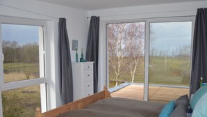 Une chambre à coucher avec un lit en bois, une commode et une vue sur un champ verdoyant, visible à travers de grandes fenêtres.