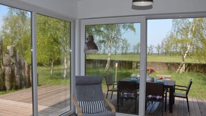 Une chambre moderne dotée d’une grande porte-fenêtre coulissante donnant sur une terrasse extérieure. L’intérieur comprend un fauteuil confortable et une table avec du mobilier d’extérieur.