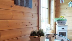 L’intérieur d’un chalet en bois, avec une table ronde, deux chaises en osier dotées de coussins à carreaux, un meuble en bois orné d’une plante et une nichoir accroché au mur.