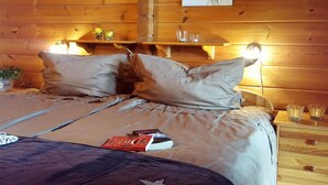 Une chambre à coucher dans une cabane en bois, avec un lit, des oreillers, une table de chevet sur laquelle sont posés des lunettes, et deux tableaux accrochés au mur.