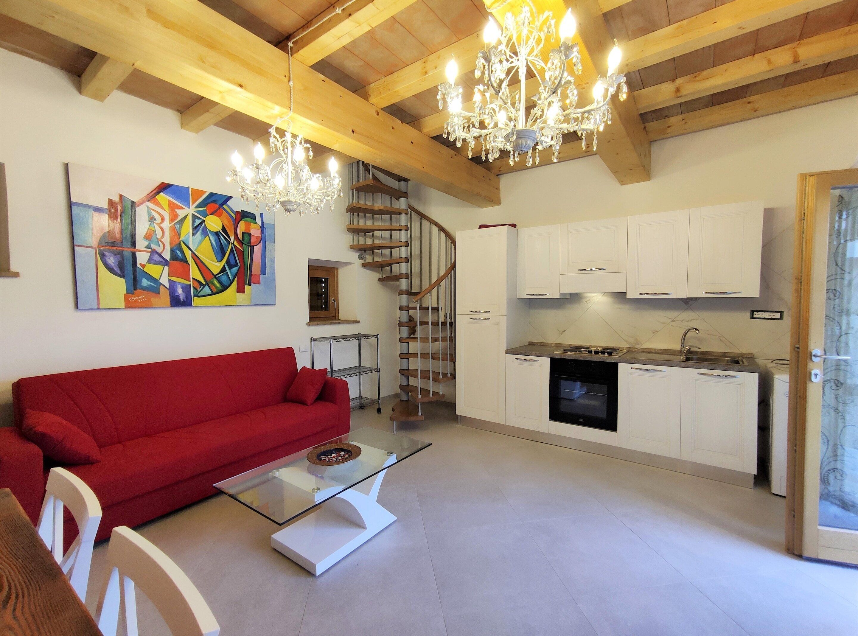 A modern living room with a red sofa, a glass coffee table, a chandelier, a staircase, and a kitchen area with white cabinets.