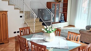 Un coin repas avec une table ronde, des chaises en bois et un vase de fleurs. Un escalier avec une rampe mène à un autre niveau.