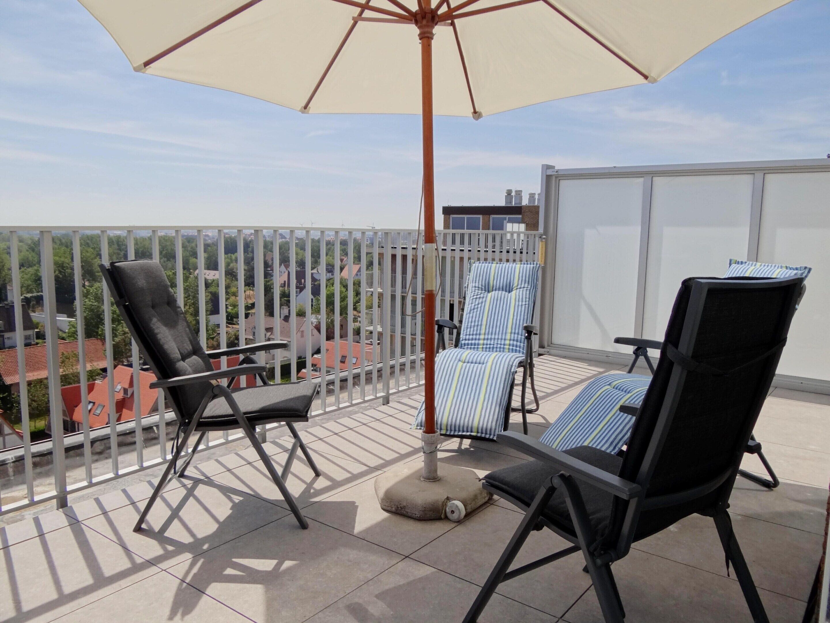 A balcony with two black lounge chairs and a striped chair, a white umbrella, and a view of buildings.