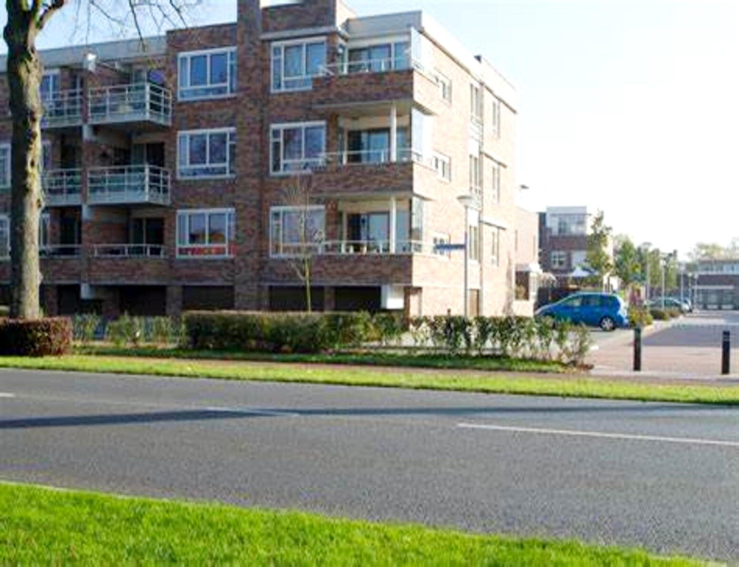 Un immeuble résidentiel à plusieurs étages, doté de balcons, d’un espace de stationnement et d’une voiture bleue garée à proximité.