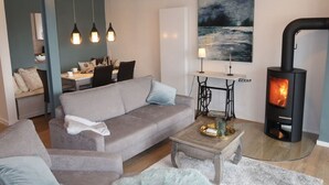 A modern living room with a sofa, a coffee table, a painting, a table lamp, pendant lights, and a wood-burning stove. - See.eck Timmel Modern Retreat (Grosefehn)
