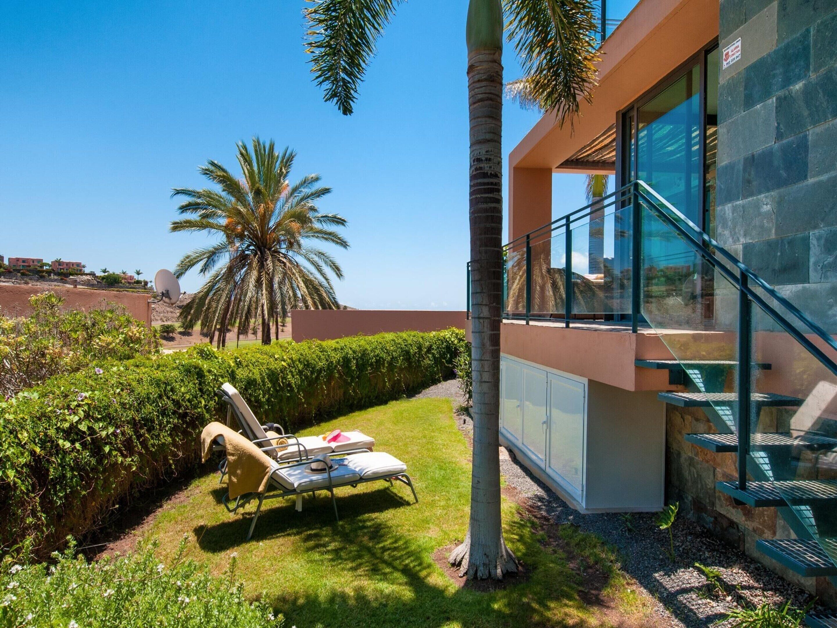 A modern building with a glass balcony, a palm tree, and a lounge area with sun loungers.