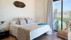 A bedroom with a bed, pillows, a bedside table, a lamp, a large window with a view, and a woven chair. - Villa in Salobre Golf With Private Pool (San Bartolomé de Tirajana)