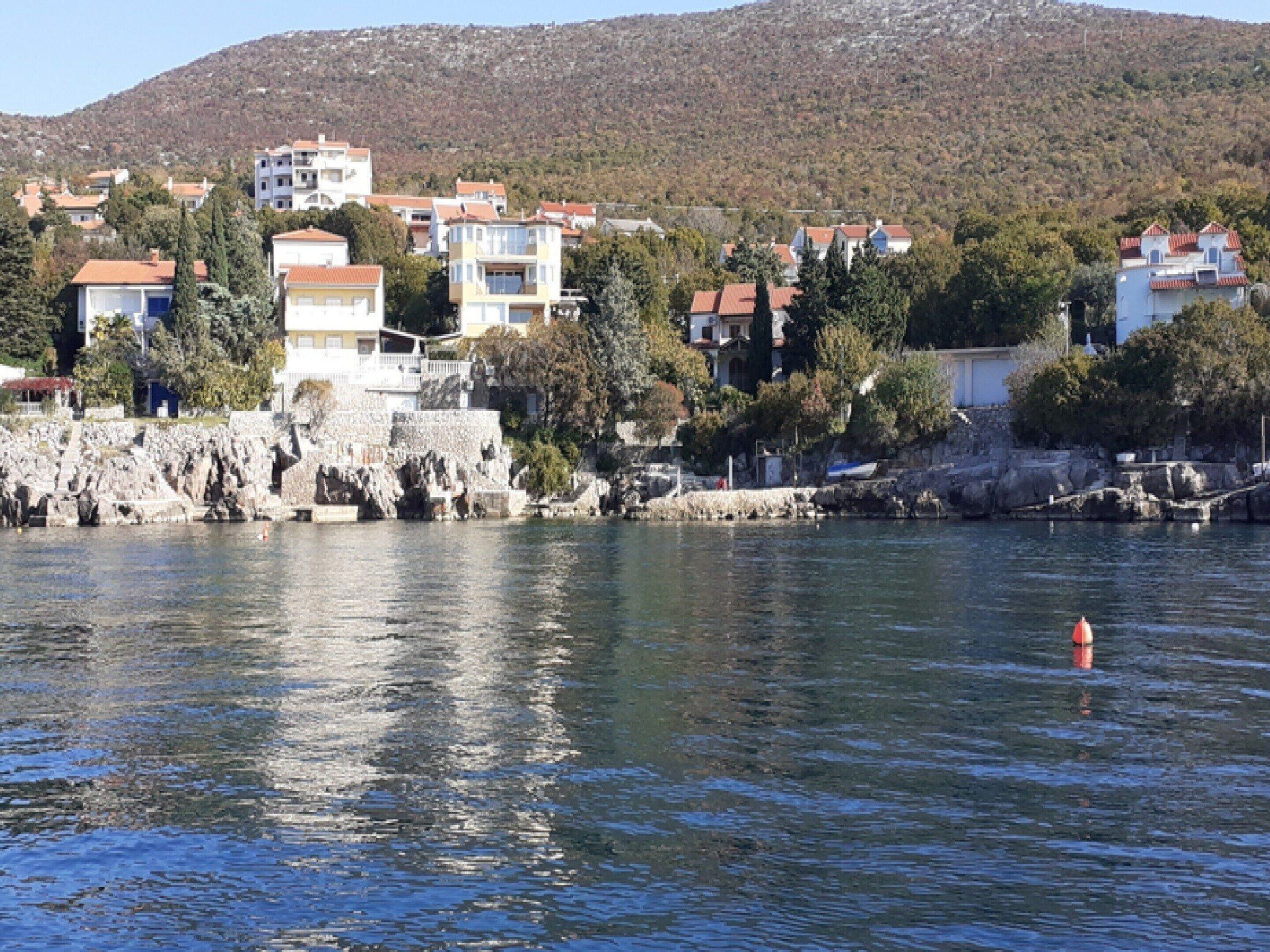 Un pueblo costero con edificios a lo largo del paseo marítimo, un litoral rocoso y una colina al fondo.