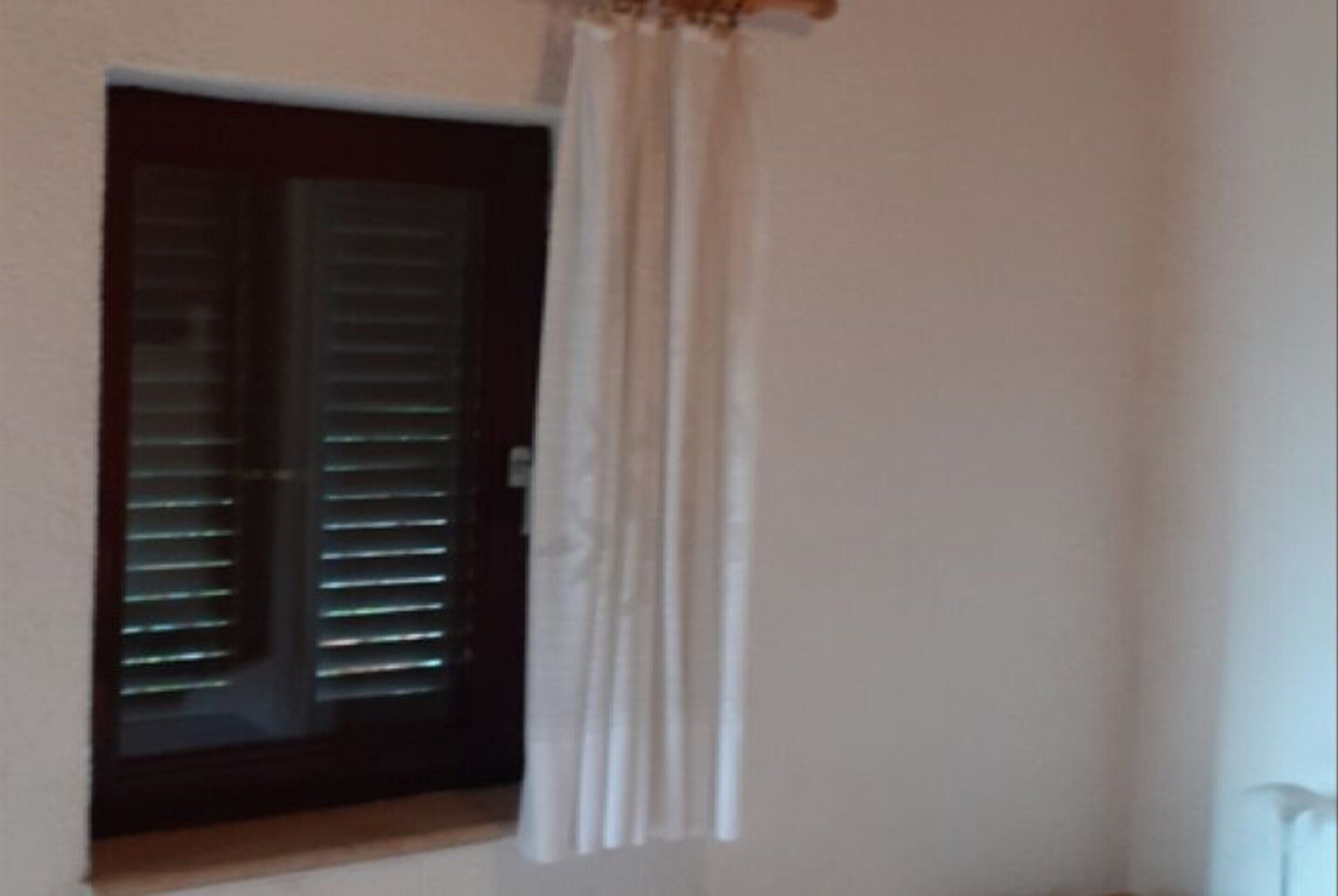 Una habitación con ventanal, persianas horizontales, una cortina blanca y una puerta con marco oscuro.
