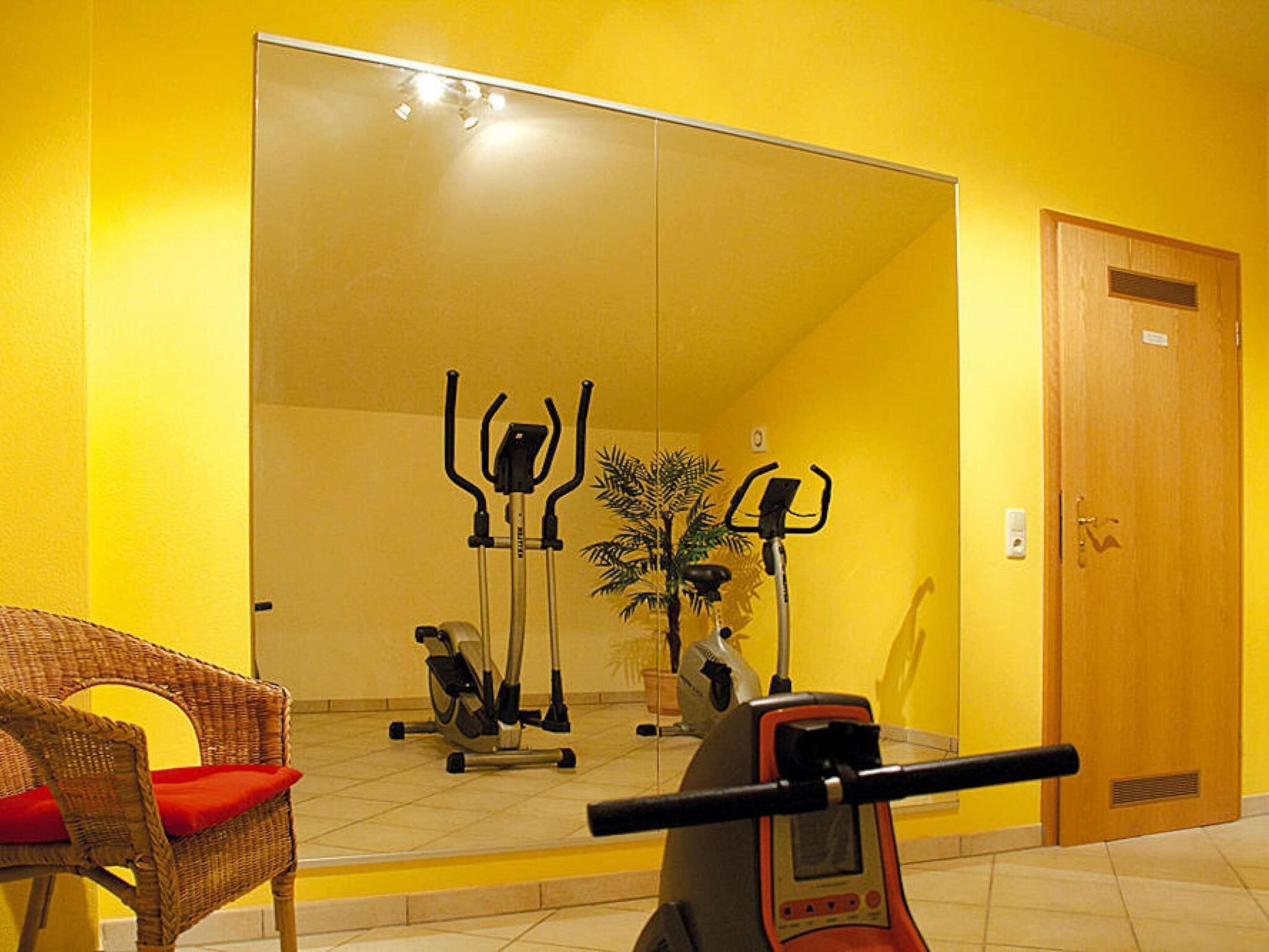 A room with a large mirror reflecting exercise equipment, a wicker chair, and a door.
