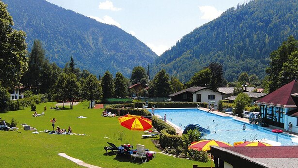 Una piscina rodeada de césped con sillas de descanso y sombrillas, con montañas al fondo.