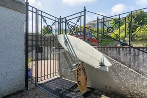 Een afgesloten gebied met een surfboard tegen een muur, een metalen poort en een trap die naar een woongebouw leidt.
