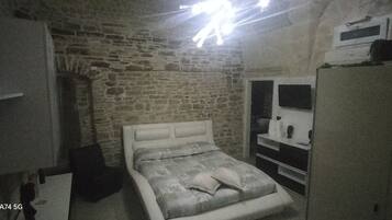 A bedroom with a stone wall, a bed with a patterned bedspread, a wardrobe, and a mirror.