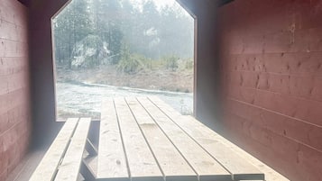 An open-sided wooden structure with a picnic table in the center, surrounded by red walls, and a view of a forested area with a river in the background.