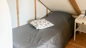 Une petite chambre avec un lit simple, un plafond en pente, un bureau en bois et un matelas gris.