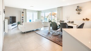 Ein modernes Wohnzimmer mit einer Couch, einem Esstisch und Stühlen. Es gibt einen Fernseher und ein Bücherregal.
