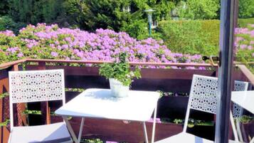 Eine Terrasse im Freien mit Tisch und zwei Stühlen, umgeben von üppigem Grün und blühenden Pflanzen.