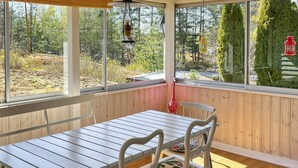 Eine überdachte Terrasse mit Holztisch und weißen Stühlen, große Fenster mit Blick auf ein bewaldetes Gebiet.