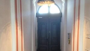 Un couloir au sol carrelé, une porte à double battant sombre au fond, et un luminaire suspendu au plafond.