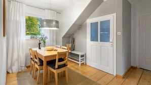 A dining area with a wooden table and chairs, a hanging light fixture, a window with curtains, and a white door with glass panels. - In het Blauwe Huis - Zolderverdieping (Garmisch-Partenkirchen)