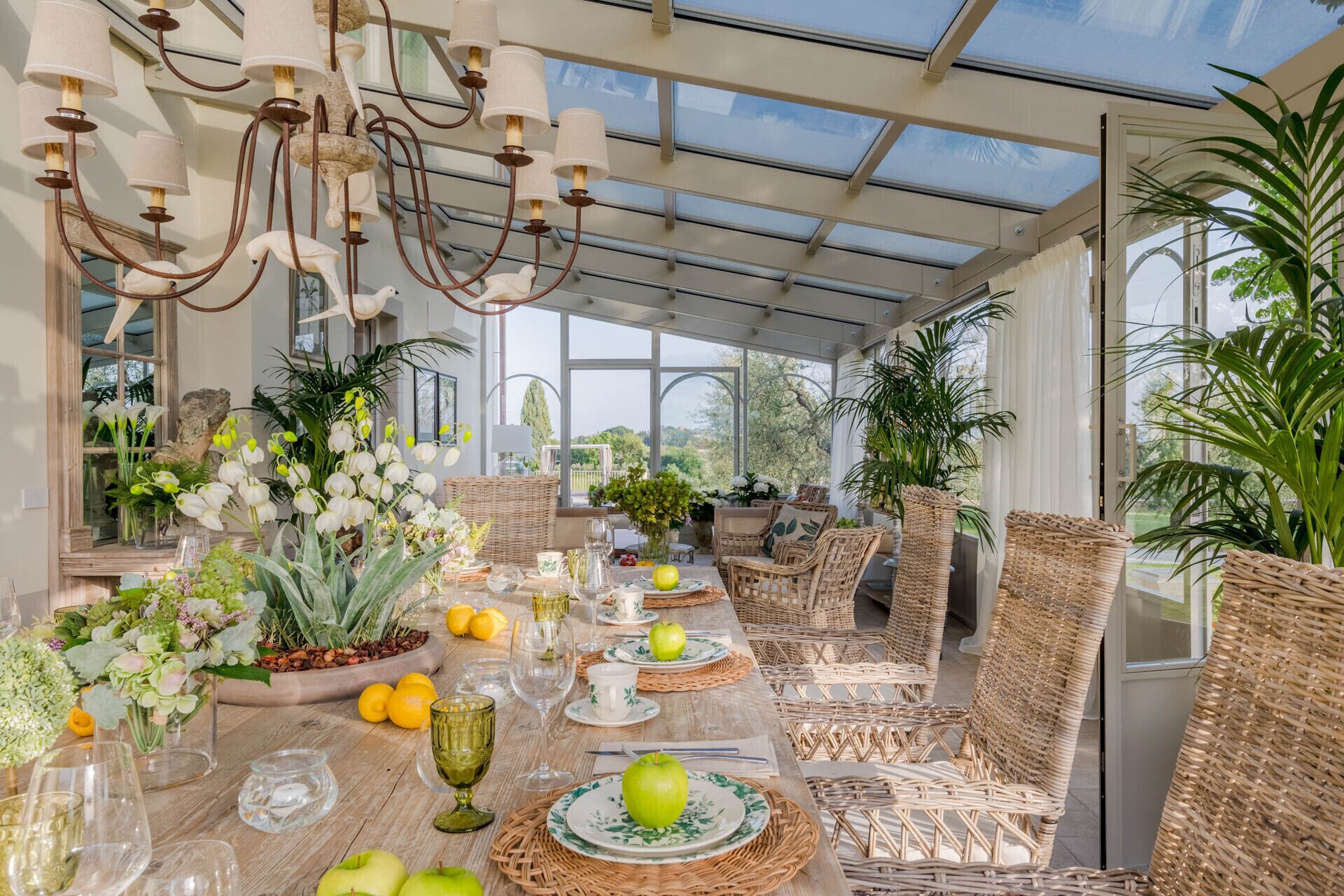 Un comedor en un invernadero con mobiliario de mimbre, techo de vidrio y varias plantas.