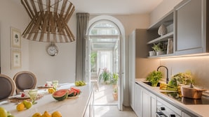 A modern kitchen with a dining area, featuring a large window, a hanging light fixture, and a variety of fruits on the counter.