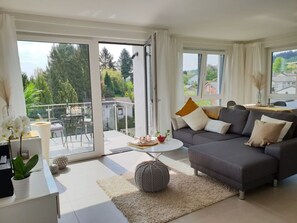 A modern living room with a grey sofa, a coffee table, and a balcony with a table and chairs. - Holiday Apartment Saartal - Senta (Merzig)
