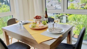 Et spisebord dækket med et udvalg af oste, druer og kringler, sammen med vinglas og en flaske vin, med udsigt over et naturskønt område med huse og grønt.