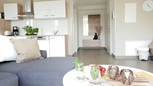 En moderne stue med en grå sofa, et rundt sofabord med glas og frugt, og et køkken i baggrunden.
