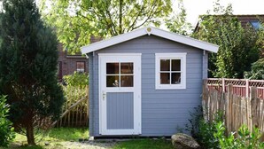 Un pequeño cobertizo de un piso con puerta y ventanas blancas, rodeado de un jardín y una valla de madera.