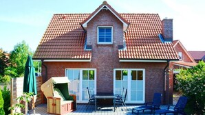 Una casa de dos pisos con techo de tejas rojas, fachada de ladrillo y una terraza de madera con mobiliario exterior.
