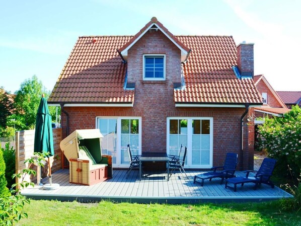 Una casa de dos pisos con techo de tejas rojas, fachada de ladrillo y una terraza de madera con mobiliario exterior.