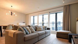 A modern living room with a large sofa, a smaller sofa, and a dining table. There is a large window with curtains and a view of buildings outside. - Barfleur Apartment With sun Terrace (De Haan)