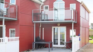 Punainen, kaksikerroksinen rakennus, jossa on parveke, lasiruutuihin jaettu ovi sekä pieni patio, jossa on pöytä ja tuolit.