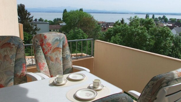 A balcony with a table set for two, overlooking a body of water and a cityscape. - With Lake and Alpine View (Immenstaad am Bodensee)