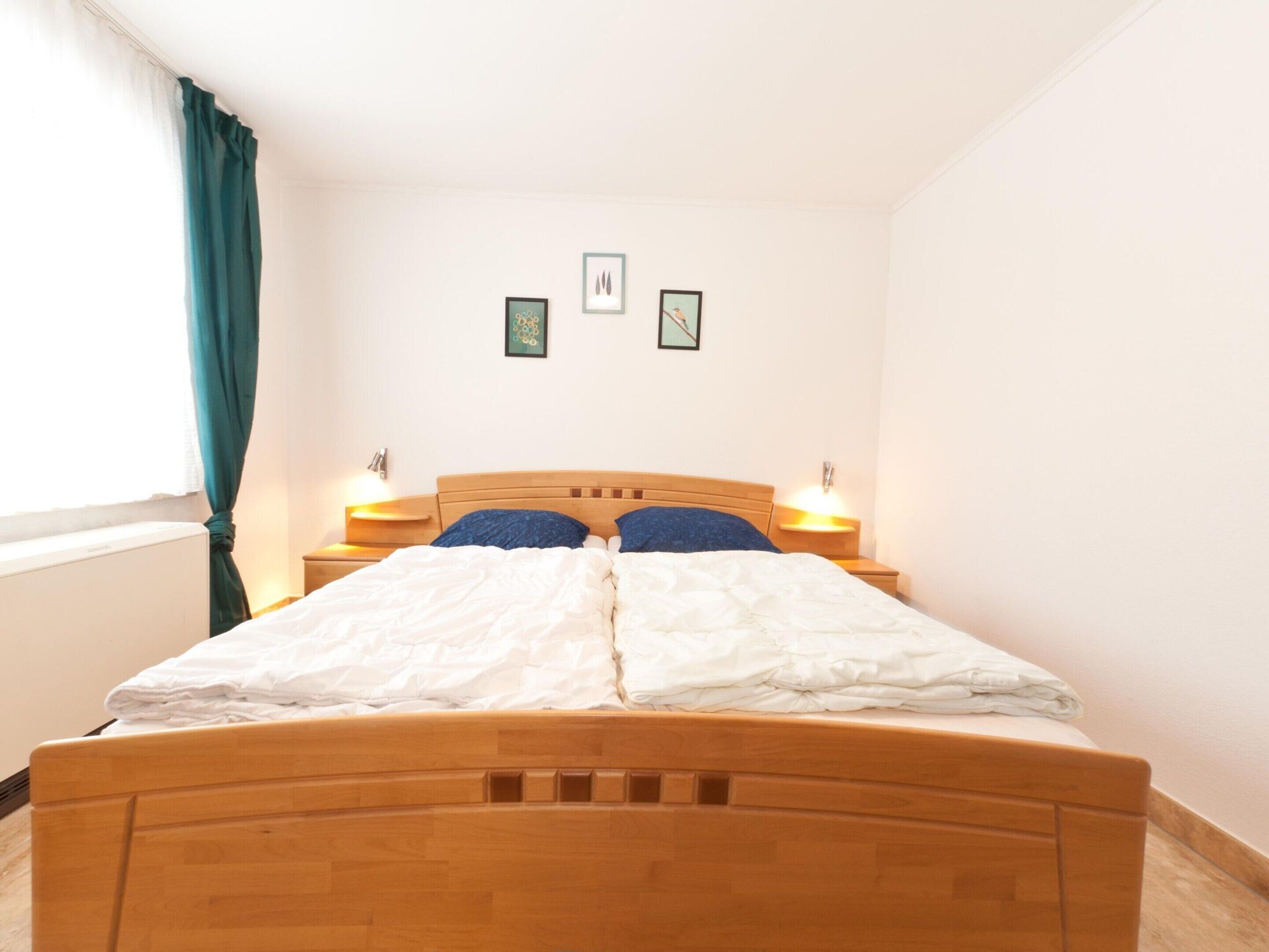 A bedroom with a wooden bed, two bedside tables with lamps, a window with curtains, and two framed pictures on the wall.