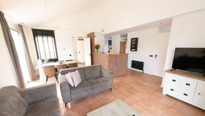 A spacious living area with a grey sofa, a coffee table, and a TV on a cabinet. There is a kitchen in the background with a dining table and chairs. - Spacious Holiday Home With Terrace (Zevenhuizen)