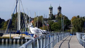 Eine Mole mit Metallgeländer, Booten und Möwen. Im Hintergrund zwei Kuppeltürme und ein Gebäude mit rotem Dach.