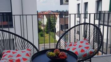 A balcony with two black chairs, a small round table with a bowl of fruit, and a view of neighboring buildings.