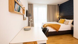 Ein kleines Hotelzimmer mit einem Bett, einem Schreibtisch, einem Bücherregal und einem Fenster mit Vorhängen.