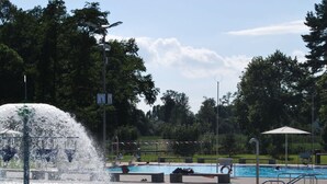 Öffentliches Schwimmbad mit einer Fontäne, umgeben von Bäumen und einer überdachten Sitzgelegenheit.