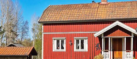 Et rødt træhus med tegltag, en veranda og et lille skur ved siden af.