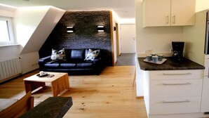 A loft-style living space with a stone accent wall, a black leather sofa, a wooden coffee table, and a kitchenette with white cabinets. - Amazing Apartment in Unterkirnach (Unterkirnach)