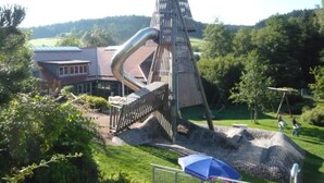 A playground with a slide and climbing structures, surrounded by greenery and a building with a red roof. - Amazing Apartment in Unterkirnach (Unterkirnach)