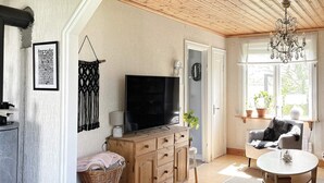 Un salon chaleureux, doté d’un téléviseur, de meubles en bois, d’un lustre et offrant une vue sur l’extérieur grâce à de grandes fenêtres.