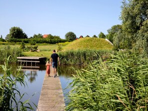 A wooden pier extending over a calm body of water, surrounded by lush greenery and a small hill in the background. - Safari Lodge Near Ijsselmeer Glamping (Andijk)